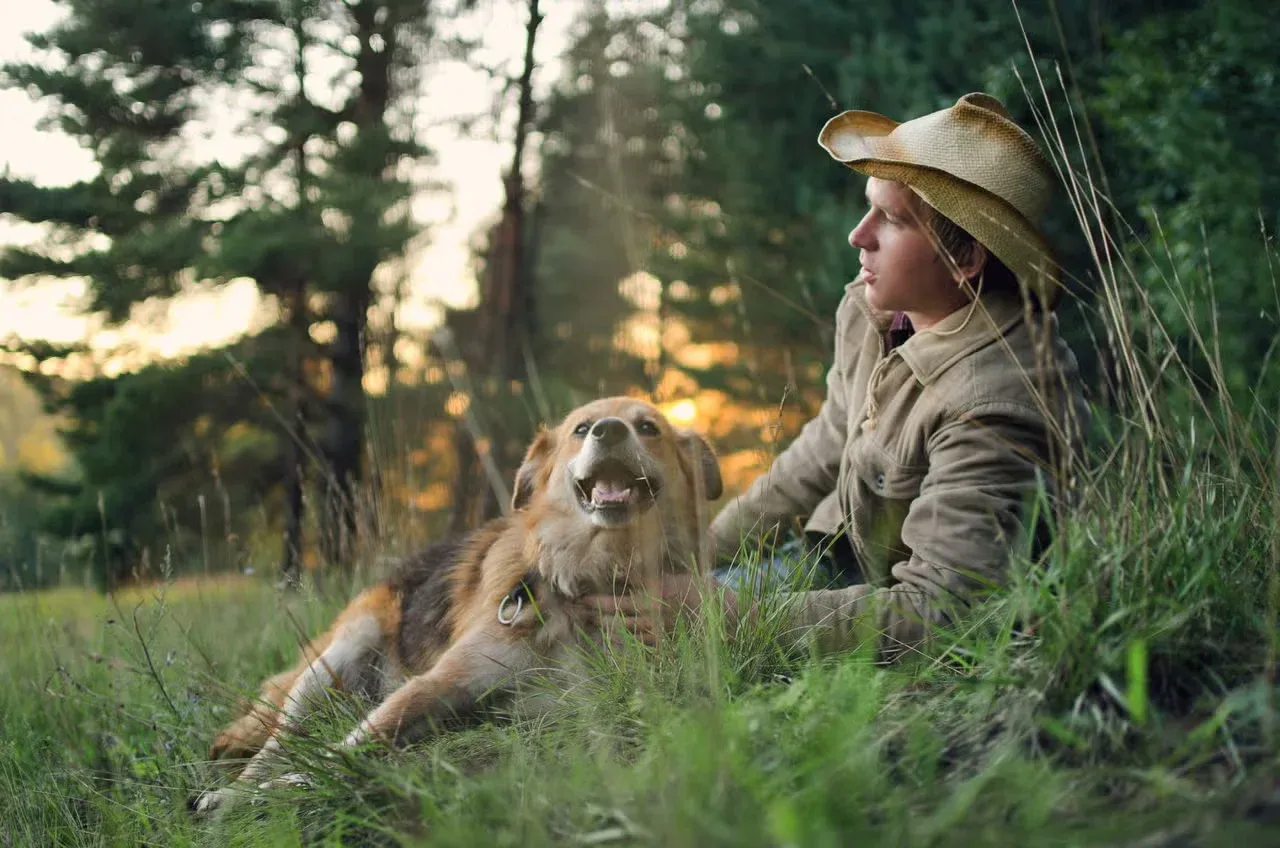 A man in a cowboy hat and his country dog relaxing outside.